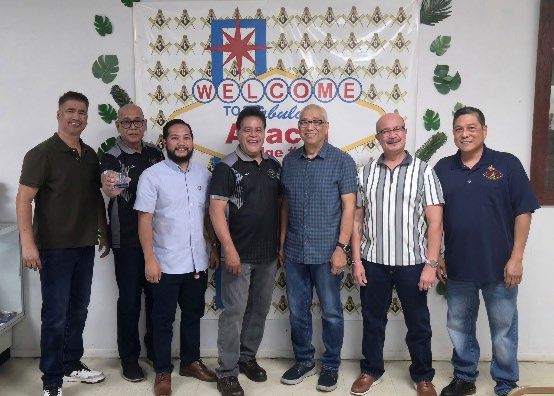 Seven Masons in casual shirts stand in a line in front of a large welcome banner decorated with square-and-compasses emblems.